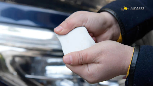 A man stretching a CarCarez Auto Detailing Magic Clay Bar before applying it to the surface of a black car. The clay bar is being prepared for use in removing contaminants and enhancing the car’s finish.