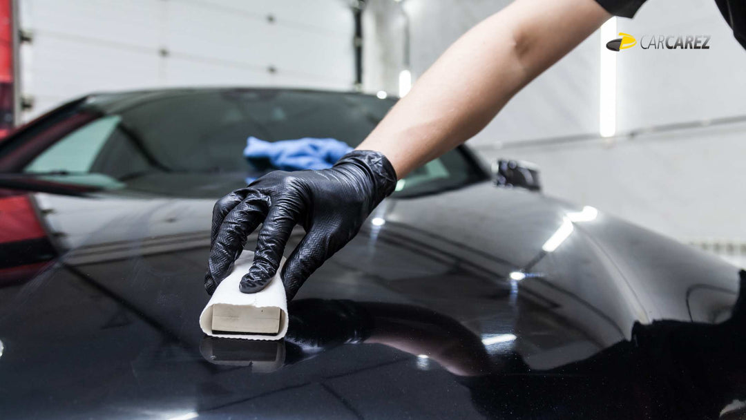 A professional car detailer wearing black gloves applies a ceramic coating to a black car’s surface using an applicator pad.