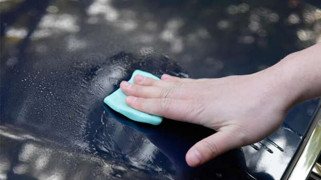 Hand rubbing a blue detailing clay bar over a wet car surface to remove contaminants.