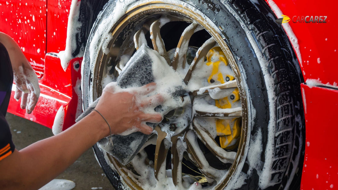 Person cleaning a car wheel with a sponge and soap, focusing on the gold alloy rim and tire, showing detailed car care during a wash.