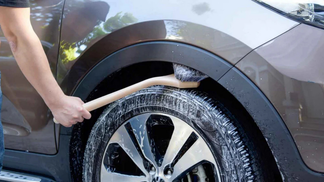 Person cleaning a car wheel using a long-handled detailing brush, with visible soap suds on the tire and rim.